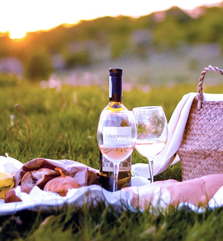 Picnic scene with wine bottle, glass, bread, and pastries on a white cloth in a grassy field at sunset. A woven bag is nearby.