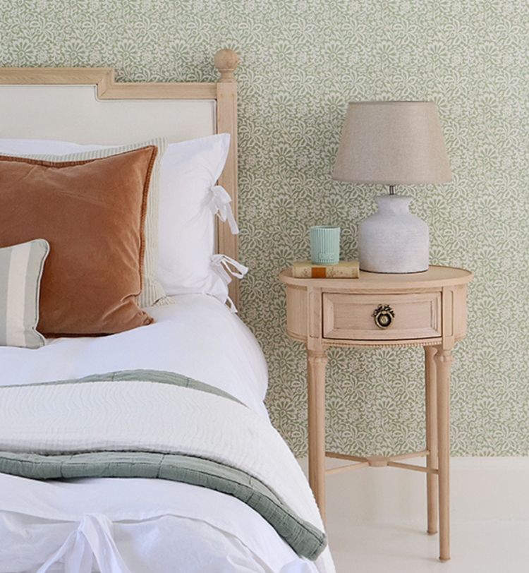 Elegant bedroom with a wooden bed, white and brown pillows, green throw, and a side table with a lamp, book, and glass against patterned wallpaper.
