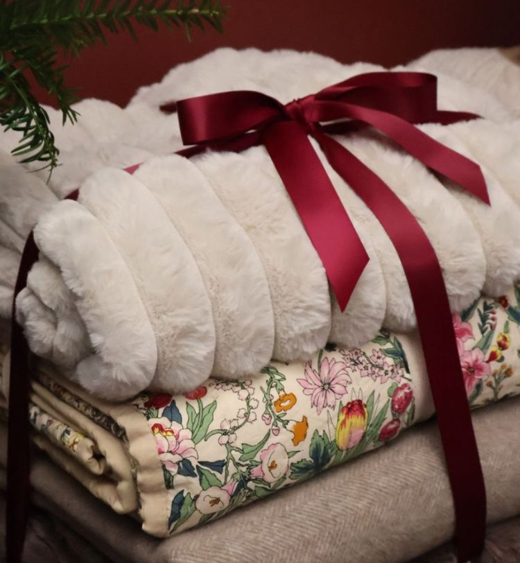 A fluffy white blanket tied with a red ribbon, resting on floral and beige blankets, with a hint of greenery in the corner.