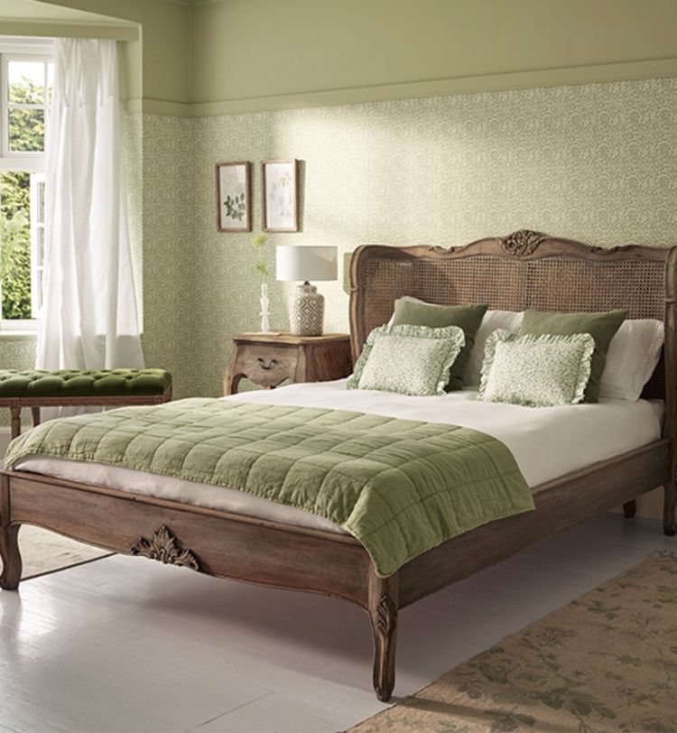 Elegant bedroom with a vintage wooden bed, green quilt, matching cushions, and floral wallpaper. Light filters through a window, illuminating the room.