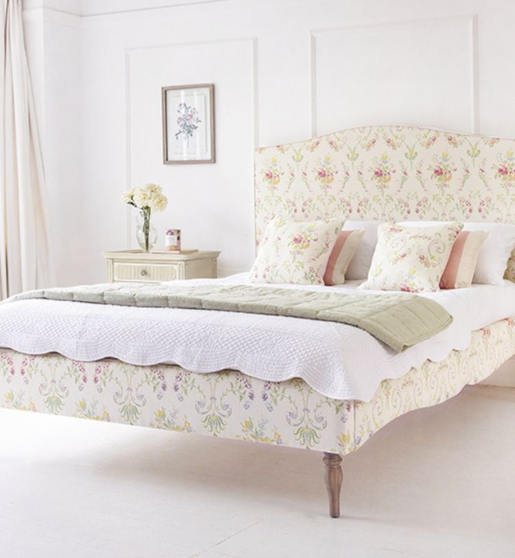 A floral-patterned bed with a green throw, white and floral pillows, beside a nightstand with flowers, in a bright, airy bedroom.