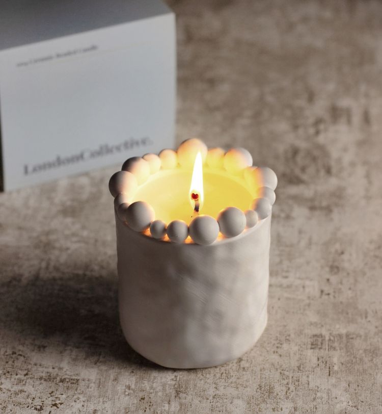 Lit candle in a white, textured holder with a beaded rim, placed on a rustic surface. A box labeled "London Collective" is in the background.