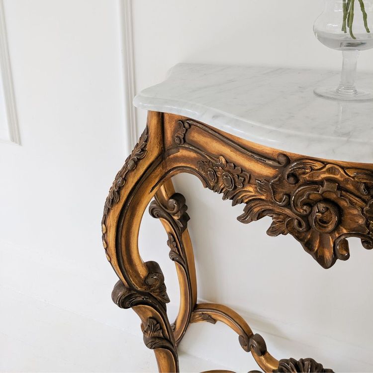 Ornate wooden console table with intricate carvings and a white marble top, set against a white paneled wall.