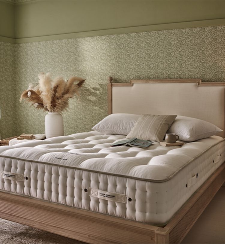 Cozy bedroom with a plush white mattress on a wooden bed frame, decorative pillows, a vase with pampas grass, and patterned wallpaper.