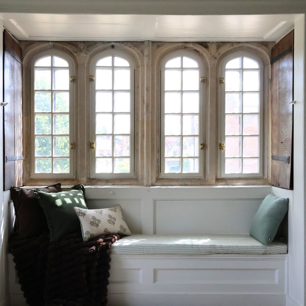 A cozy window nook with a cushioned bench, pillows, and a view through four arched, multi-paned windows framed by wooden shutters.