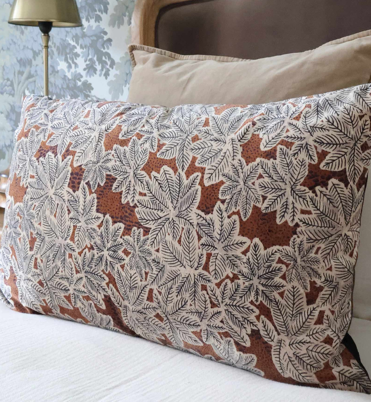 A decorative pillow with a white leaf pattern on a brown background rests against beige pillows on a bed with a floral wallpaper backdrop.
