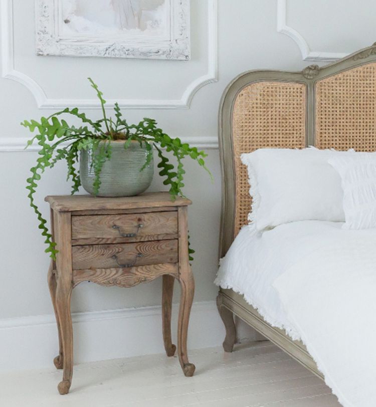 A vintage wooden bedside table with a potted fern beside a cane headboard bed with white bedding in a softly lit room.