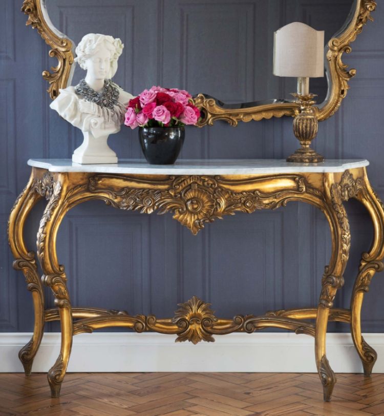 Elegant console table with ornate gold detailing, topped with a white bust, pink roses in a black vase, and a lamp, against a paneled wall.