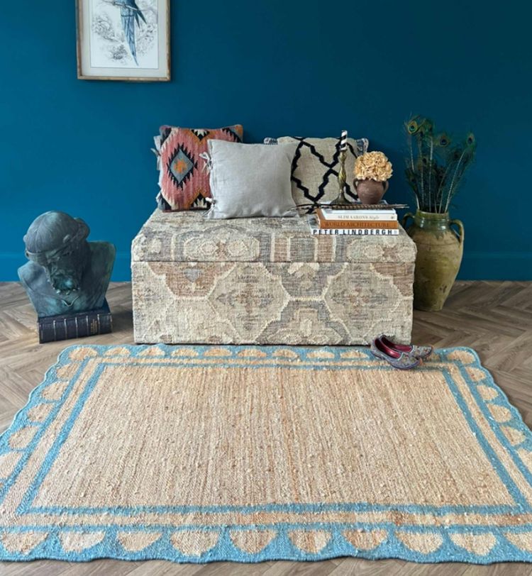 Room with a blue wall, patterned bench, decorative pillows, a bust sculpture, a large vase, and a woven rug with blue accents on wooden flooring.
