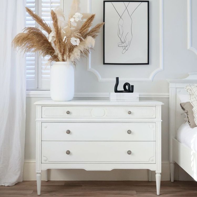 White dresser with pampas grass in a vase, abstract hand drawing above, and a decorative black sculpture. Elegant, minimalist bedroom decor.