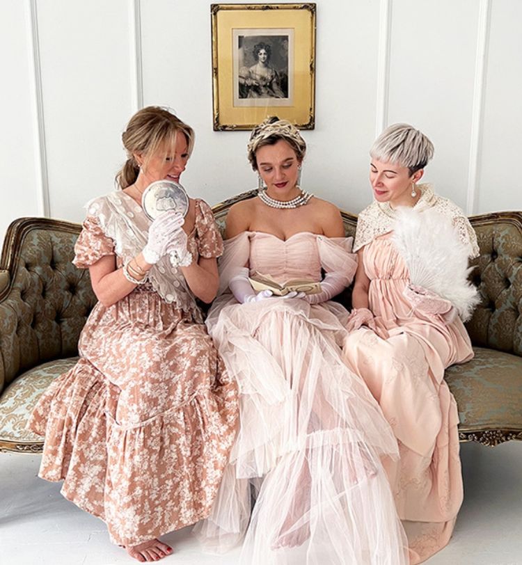 Three women in vintage dresses sit on a sofa, one holding a mirror, another a book, and the third a fan, beneath a framed portrait.