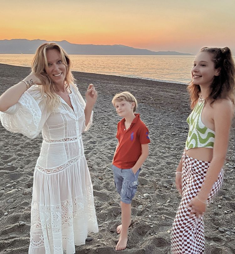 Three people on a beach at sunset: a woman in a white dress, a boy in a red shirt, and a girl in a patterned outfit.