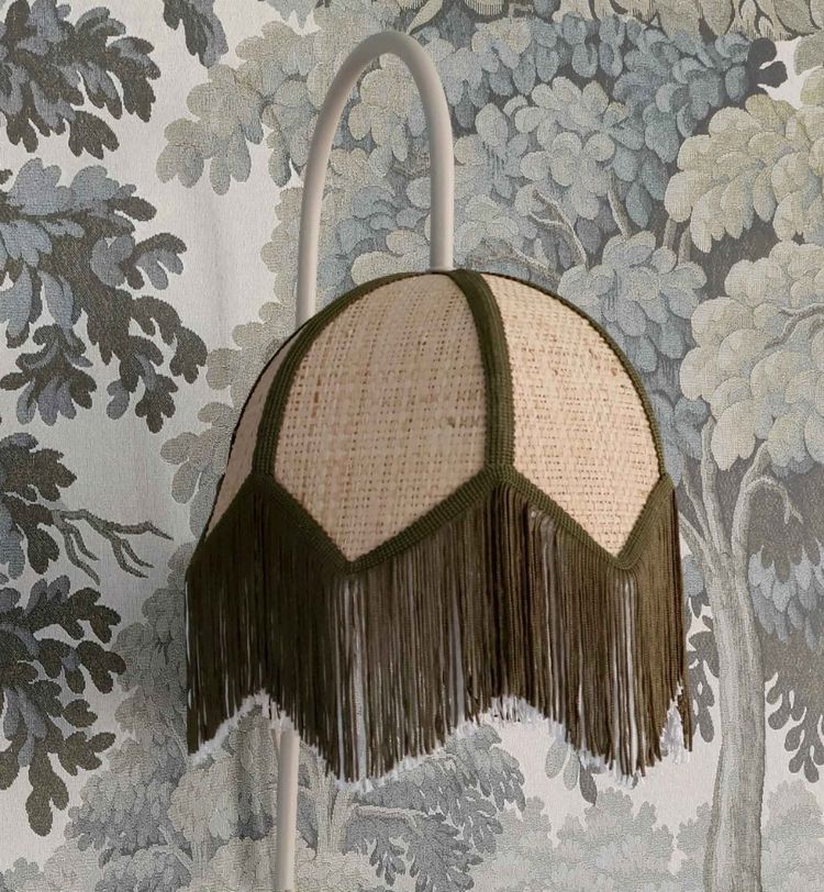 A vintage-style lampshade with fringe, set against a wallpaper featuring a detailed tree and foliage pattern in muted tones.