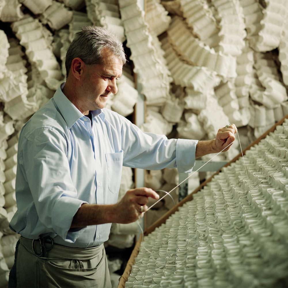 Man in a workshop carefully strings together white ceramic cups, surrounded by stacks of similar cups in the background.