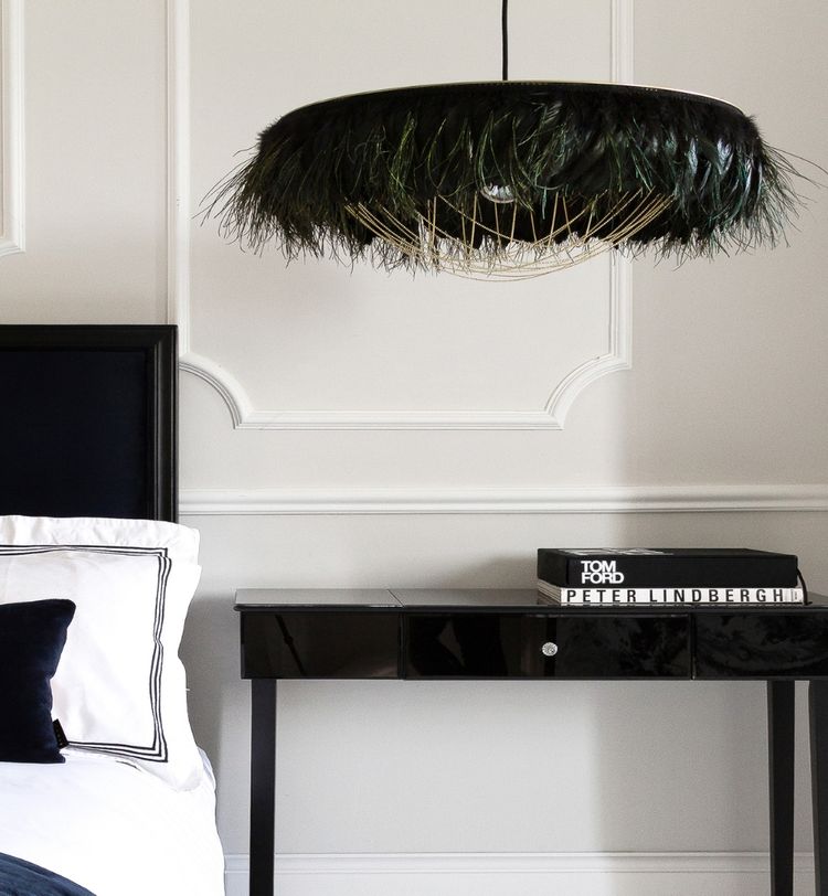 Elegant bedroom with a feathered pendant light, black side table, and books. White walls and a neatly made bed with black and white linens.