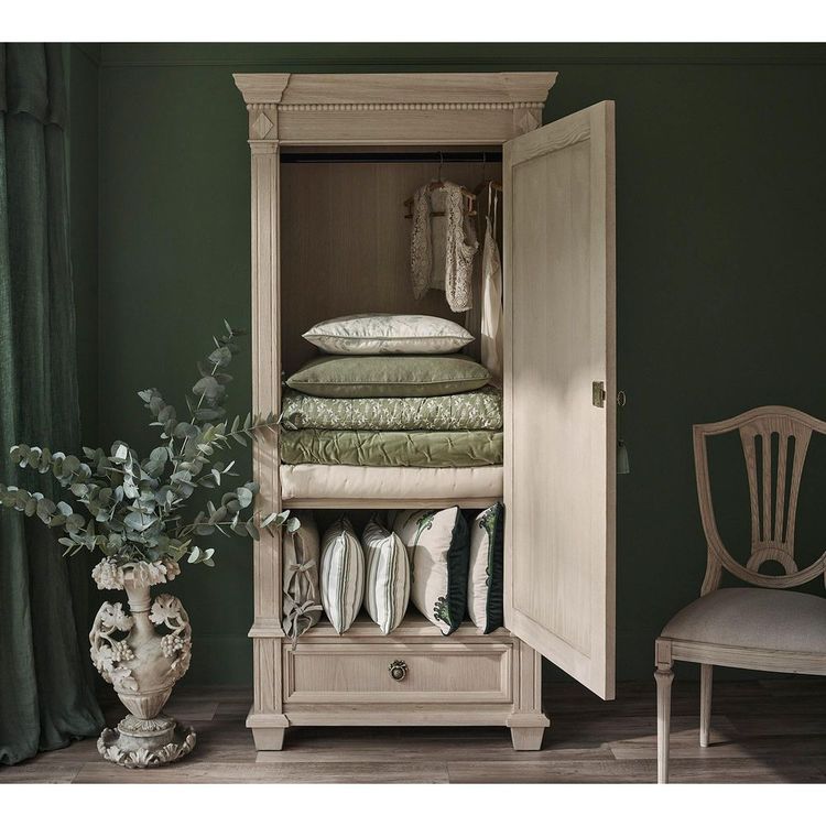 Elegant wooden wardrobe with open door, revealing neatly stacked green and white linens and pillows. Nearby, a decorative vase with greenery.