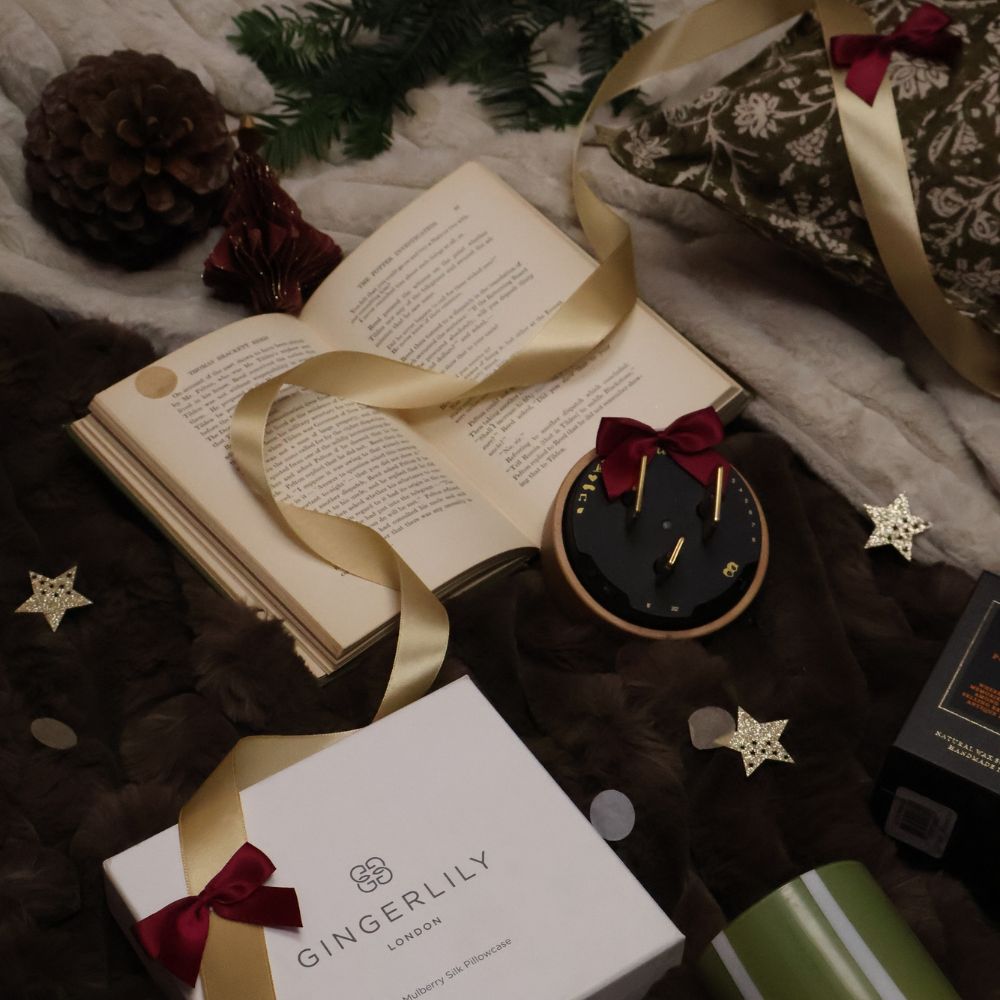 Cozy scene with an open book, ribbon, decorative clock, pinecone, and a gift box labeled "Gingerlily London" on a soft, dark surface.