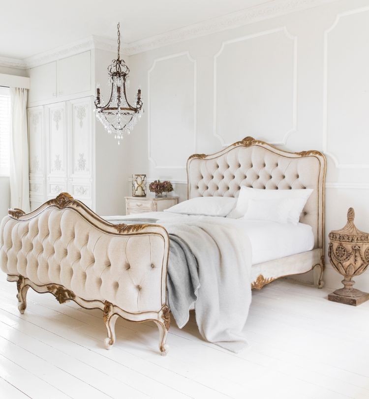 Elegant bedroom with a vintage-style tufted bed, ornate chandelier, and white decor. A plush blanket is draped over the bed.