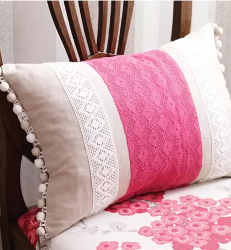 A decorative pillow with pink lace and white crochet accents, adorned with pom-poms, rests on a floral-patterned chair.