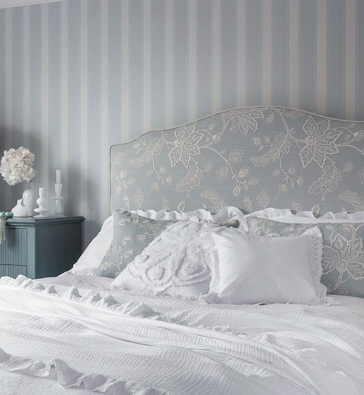 Elegant bedroom with a floral-patterned headboard, white and blue pillows, and a white bedspread. A nightstand with decor is on the left.