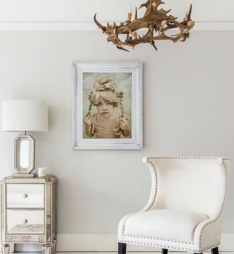 Elegant room with a white chair, mirrored side table, ornate lamp, antler chandelier, and a framed portrait of a child on the wall.