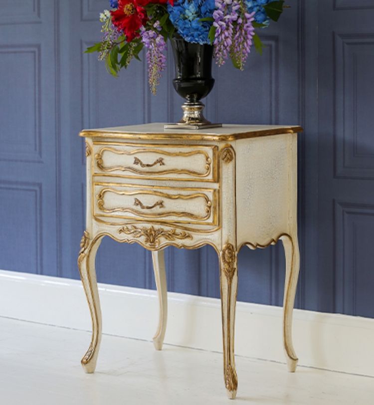 Elegant vintage side table with ornate gold trim, featuring two drawers and a vase of colorful flowers on top, set against a blue paneled wall.