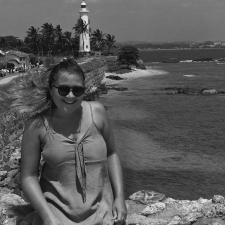 Woman in sunglasses smiling by a rocky shoreline with a distant lighthouse and palm trees in the background.