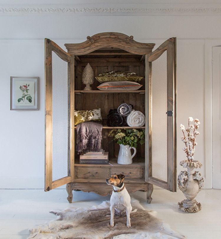 A rustic wooden cabinet with open doors displays pillows and blankets. A small dog sits on a fur rug in front, and a floral vase is nearby.