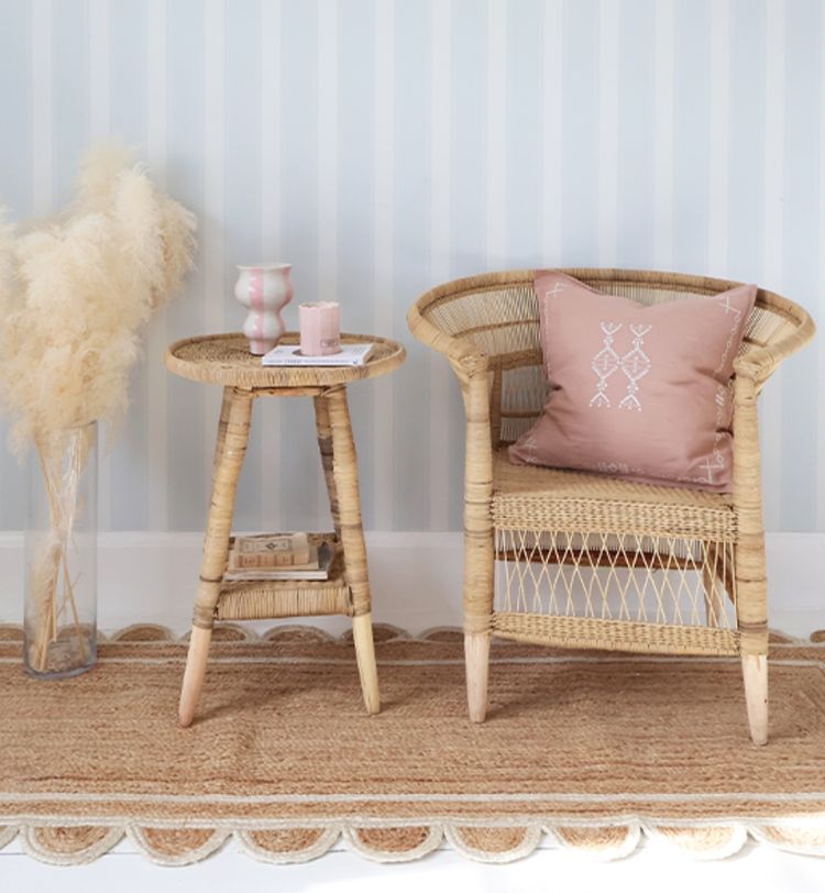 Wicker chair with a pink pillow next to a small round wicker table holding a candle and vase, set on a woven rug with pampas grass in a vase nearby.