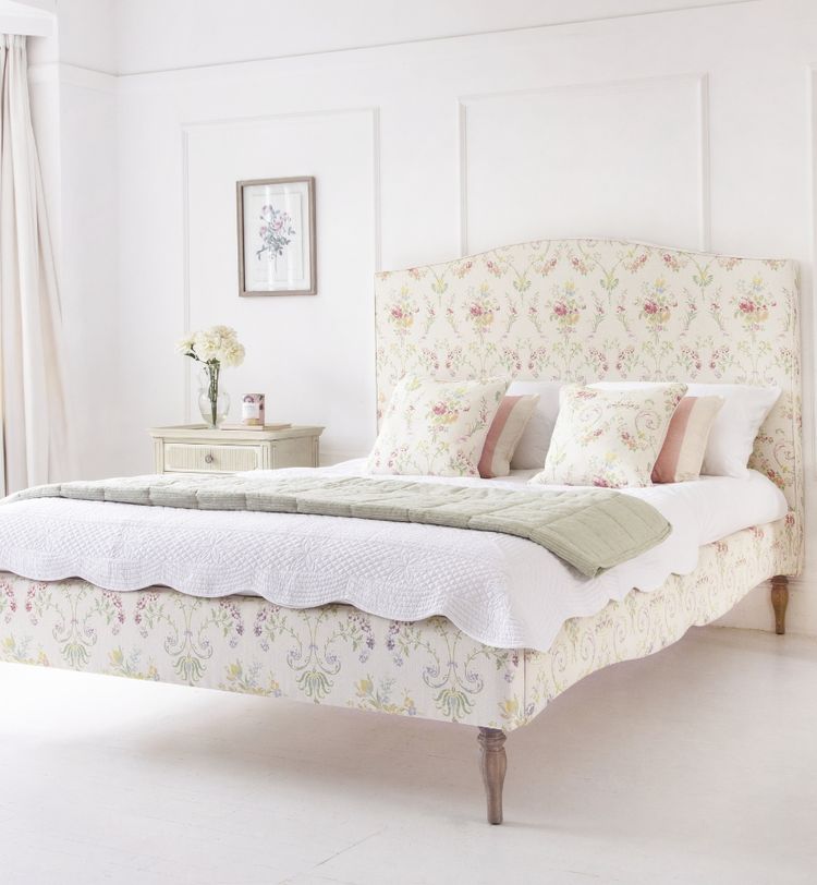 Elegant bedroom with a floral-patterned bed, white bedding, and pastel pillows. A small nightstand with flowers and a framed picture adorn the space.
