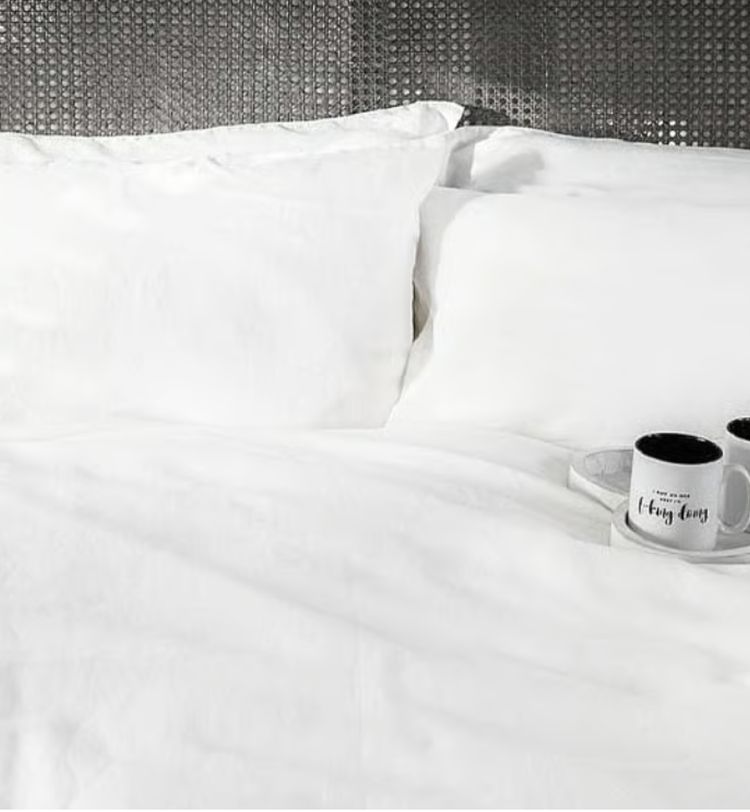 White bed with two pillows and two mugs on a tray, placed on a textured gray headboard background.
