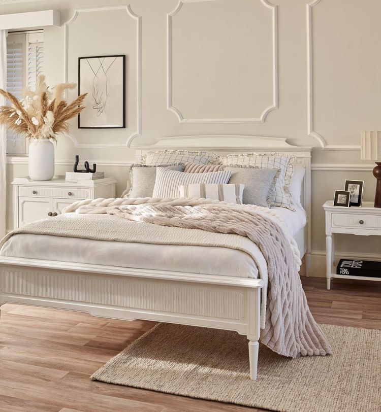 Elegant bedroom with a white bed, textured pillows, a beige knit throw, nightstands, a lamp, pampas grass, and wall art on a wooden floor.