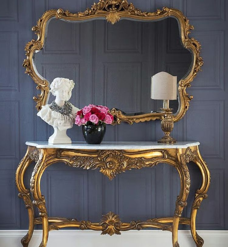 Ornate gold console table with a large mirror, topped with a white bust, a vase of pink roses, and a small lamp, against a dark paneled wall.
