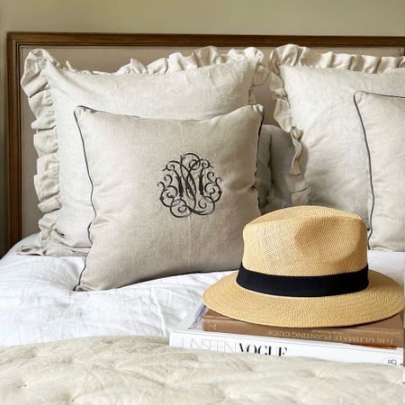 A neatly made bed with ruffled beige pillows, a monogrammed cushion, a straw hat, and stacked fashion books on a white duvet.