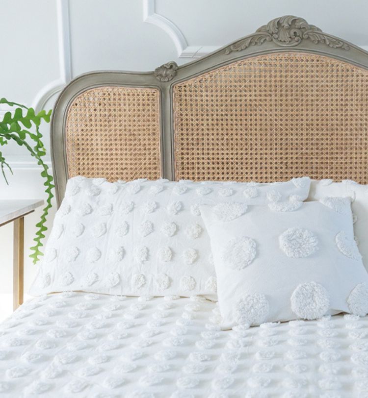 A cozy bed with a wicker headboard, white textured bedding, and a decorative pillow. A green plant sits on a bedside table.