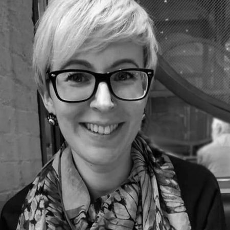 A person with short hair, glasses, and a patterned scarf smiles while sitting indoors, with a brick wall and window in the background.
