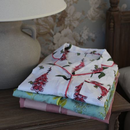 Folded floral pajamas stacked on a wooden nightstand beside a lamp, with a floral-patterned wallpaper in the background.