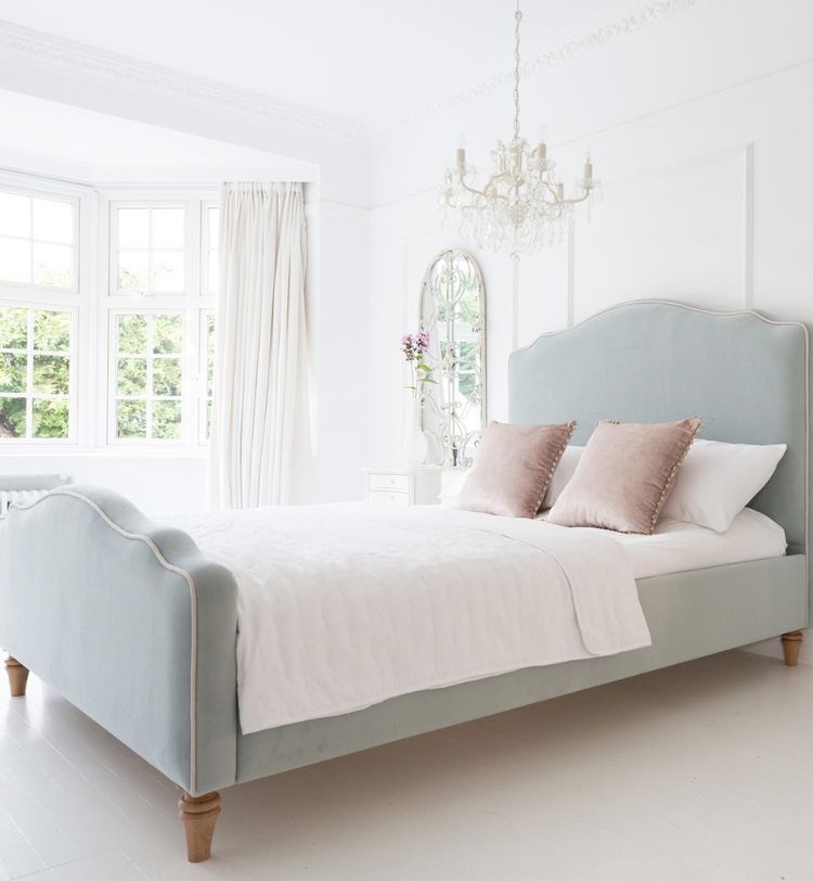 Elegant bedroom with a light blue upholstered bed, white bedding, pink pillows, a chandelier, and a large window with sheer curtains.