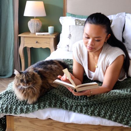 Woman reading a book on a bed with a large, fluffy cat beside her. The room has a cozy, warm ambiance with a lamp on a wooden nightstand.