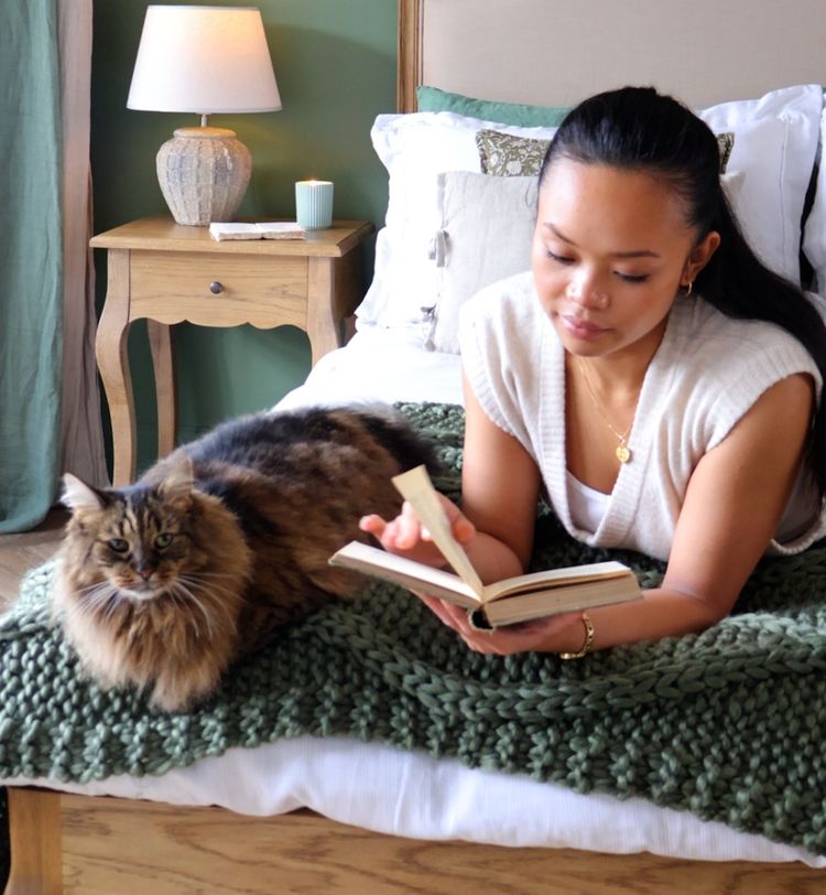 Woman reading a book on a bed with a large, fluffy cat beside her. The room has a cozy, warm ambiance with a lamp on a wooden nightstand.