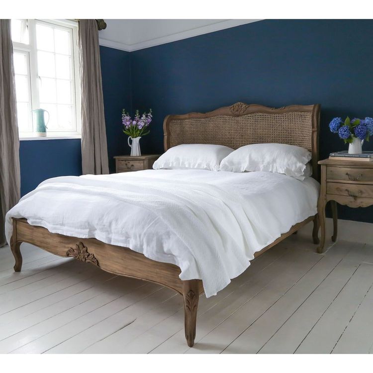Elegant bedroom with a wooden bed, white bedding, blue walls, and floral arrangements on vintage nightstands by a window.