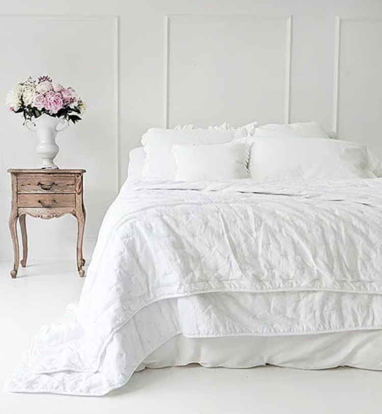 A white bed with fluffy pillows and a quilt, next to a wooden bedside table with a vase of pink and white flowers, in a bright room.