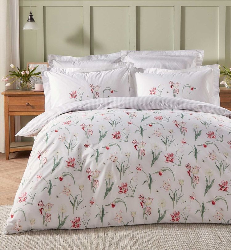 A neatly made bed with floral-patterned bedding, striped pillows, and wooden side tables with flower arrangements in a softly lit room.