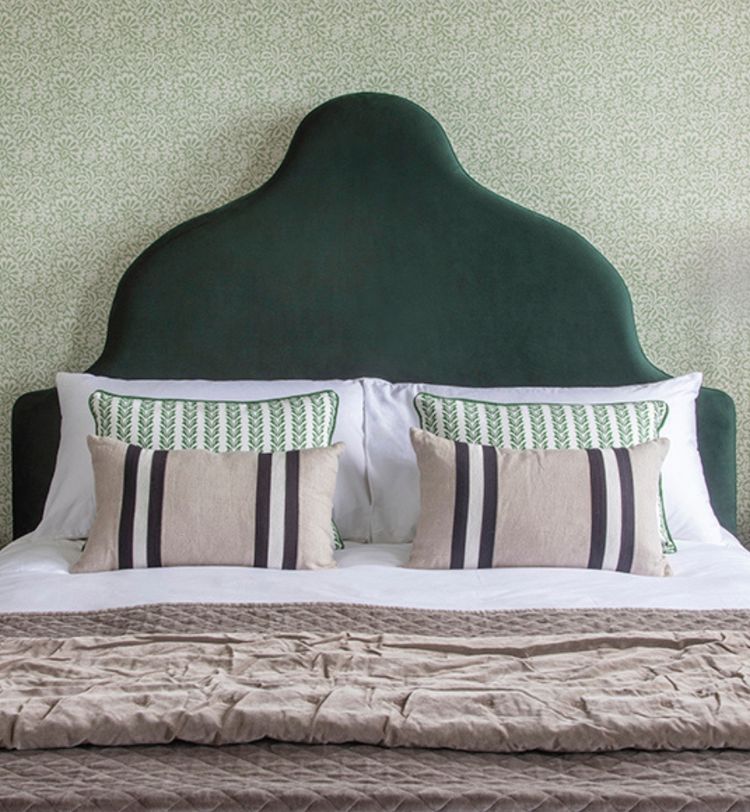 A bed with a dark green, uniquely shaped headboard, patterned pillows, and a beige quilt, set against a patterned green wallpaper.