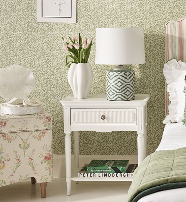 Bedroom with a white nightstand, geometric lamp, vase of pink tulips, floral chair, and green patterned wallpaper.