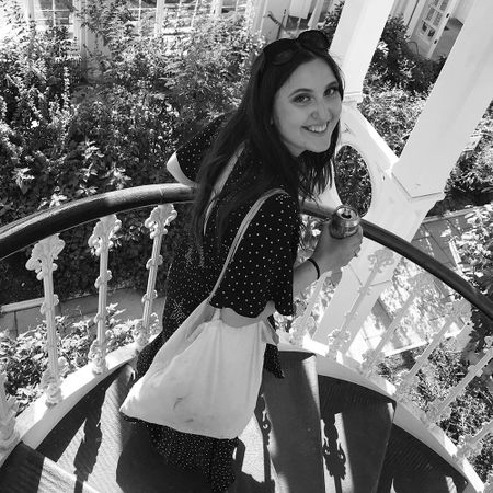 Woman in polka dot dress smiles while holding a can and climbing a spiral staircase, surrounded by lush greenery.