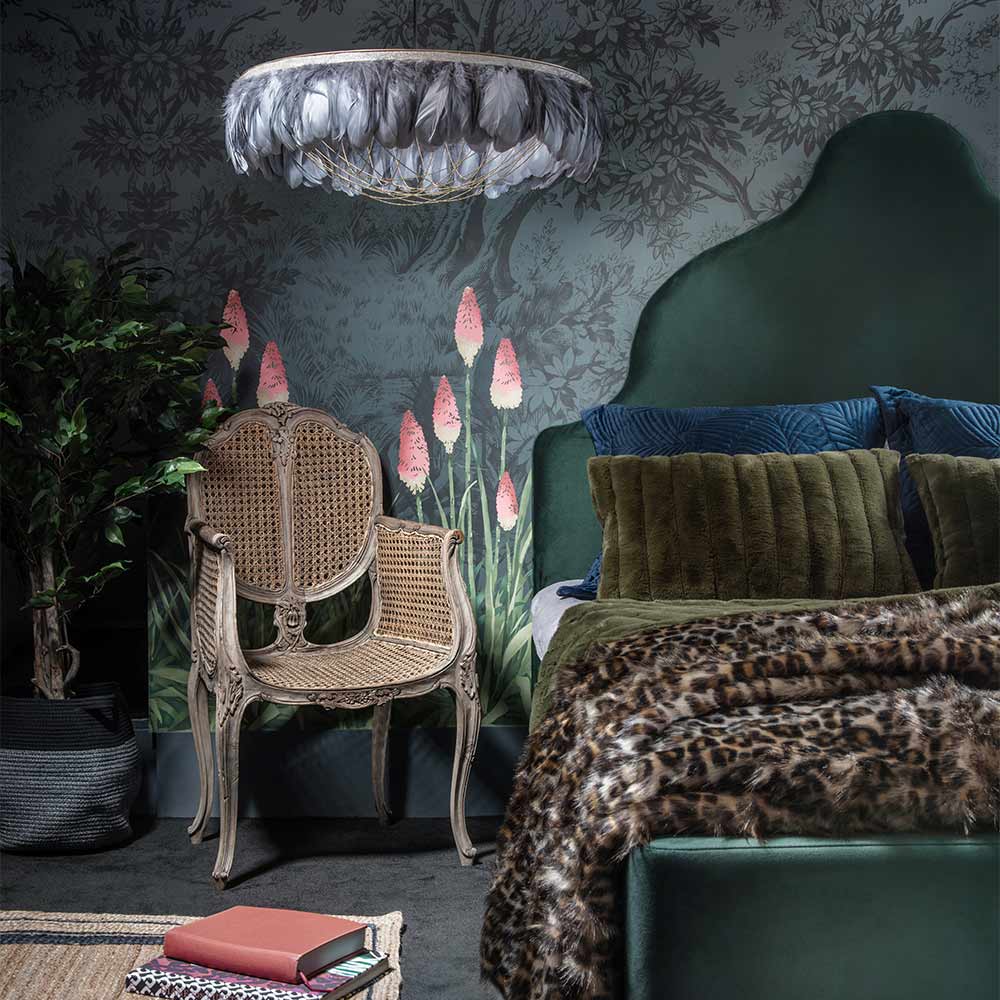 Elegant bedroom with a green velvet bed, leopard print throw, wicker chair, feather chandelier, and floral wallpaper.