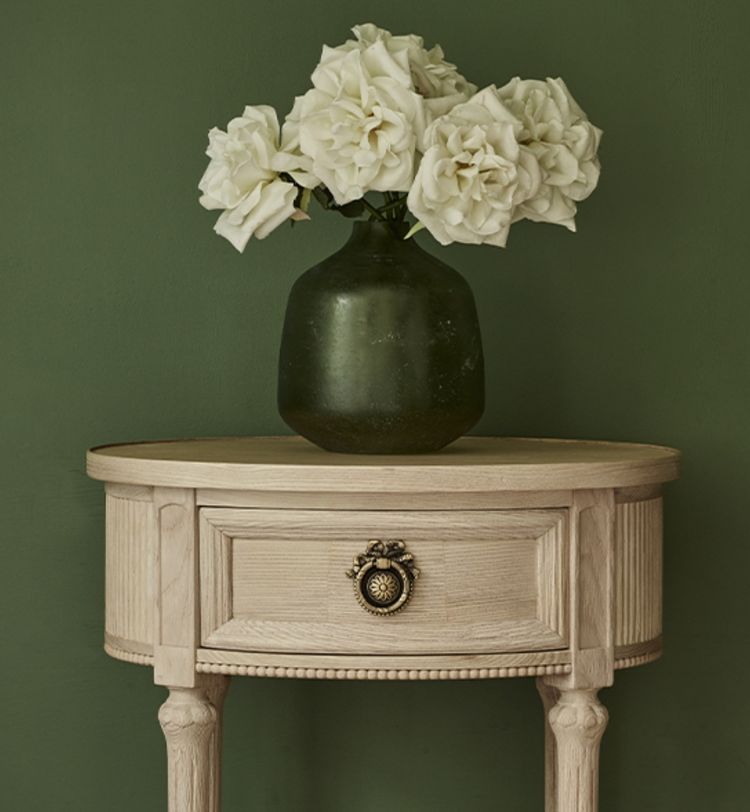Wooden table with a single drawer and ornate handle, topped with a green vase holding white roses, set against a green wall.