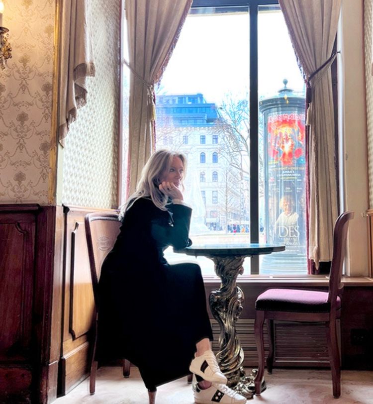 A woman sits at a small table in a cozy, elegant room with patterned wallpaper, gazing out a large window overlooking a city street.