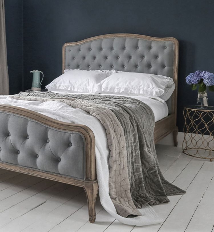 Elegant bedroom with a tufted gray bed, white and gray bedding, dark blue wall, and a side table with purple flowers in a vase.